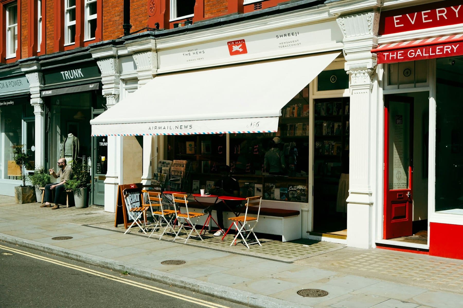 Independent shopfronts in Marylebone: a newsagent with a striped awning, a menswear shop and a gallery, with tables on the pavement.