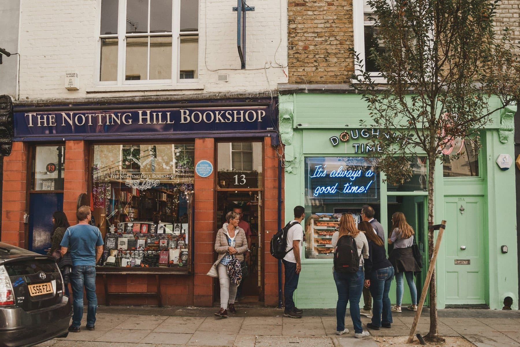 The Notting Hill Bookshop with its navy fascia, beside a mint-green doughnut shop, both with people outside.